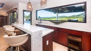 Une cuisine moderne dotée d’un grand îlot central, de tabourets de bar, de luminaires suspendus et d’une grande fenêtre offrant une vue sur un paysage.