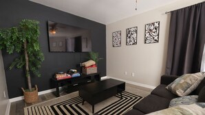 A modern living room with a black sofa, a black coffee table, a flat-screen TV, and a large plant. - North Hills/Gym/Sauna/Massage Chair/Pool (Raleigh)