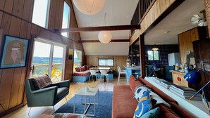 Amplia sala de estar con paredes de madera, un ventanal grande y vistas al exterior. Hay un comedor con mesa y sillas, y una cocina con armarios y electrodomésticos.