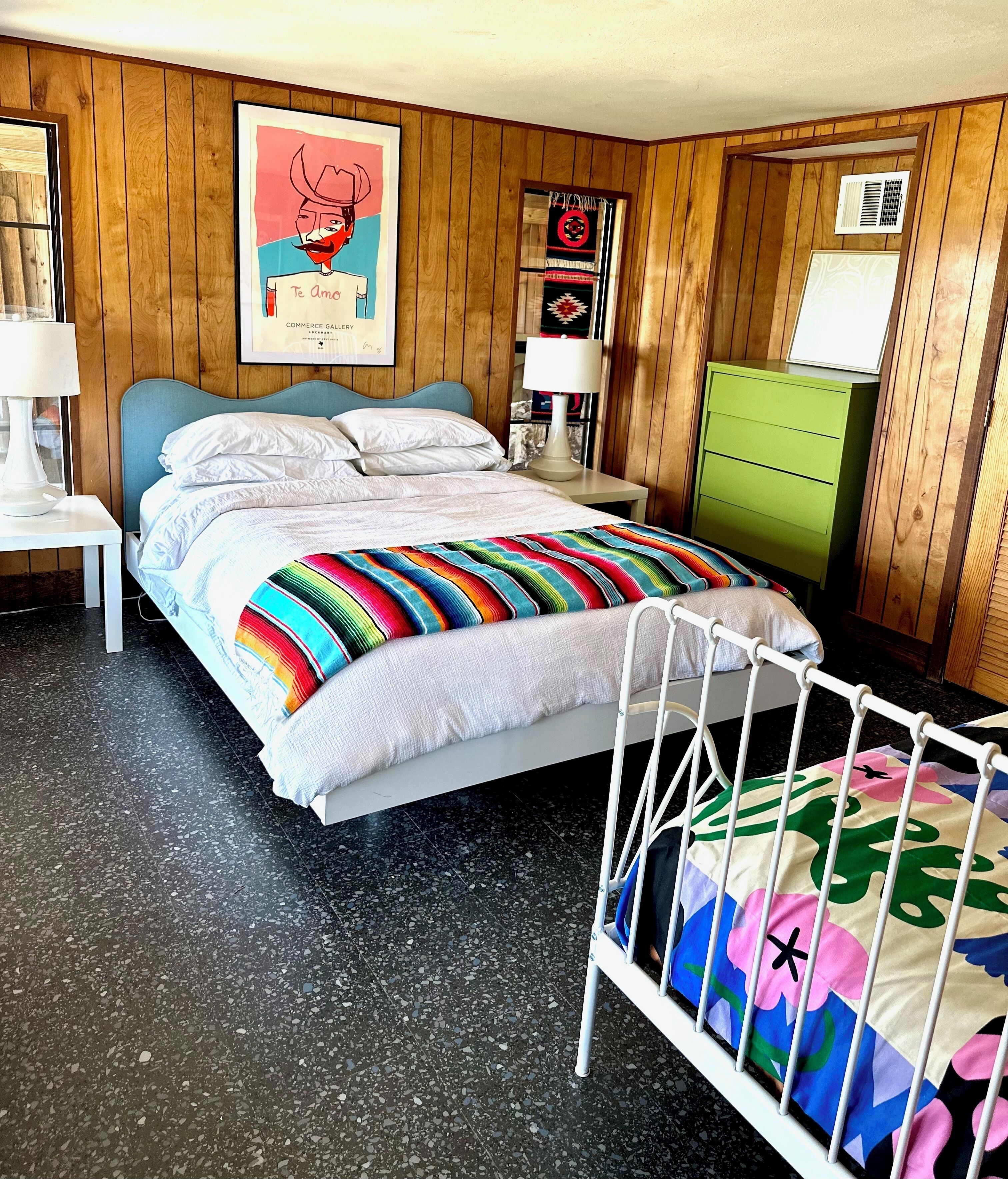 A bedroom with a bed, a crib, a green dresser, and a wall with wooden paneling.