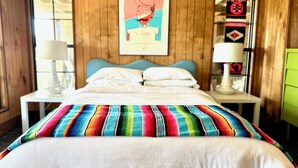 Un dormitorio con una cama, dos mesitas de noche, una lámpara de pared, un cuadro enmarcado y una manta colorida.
