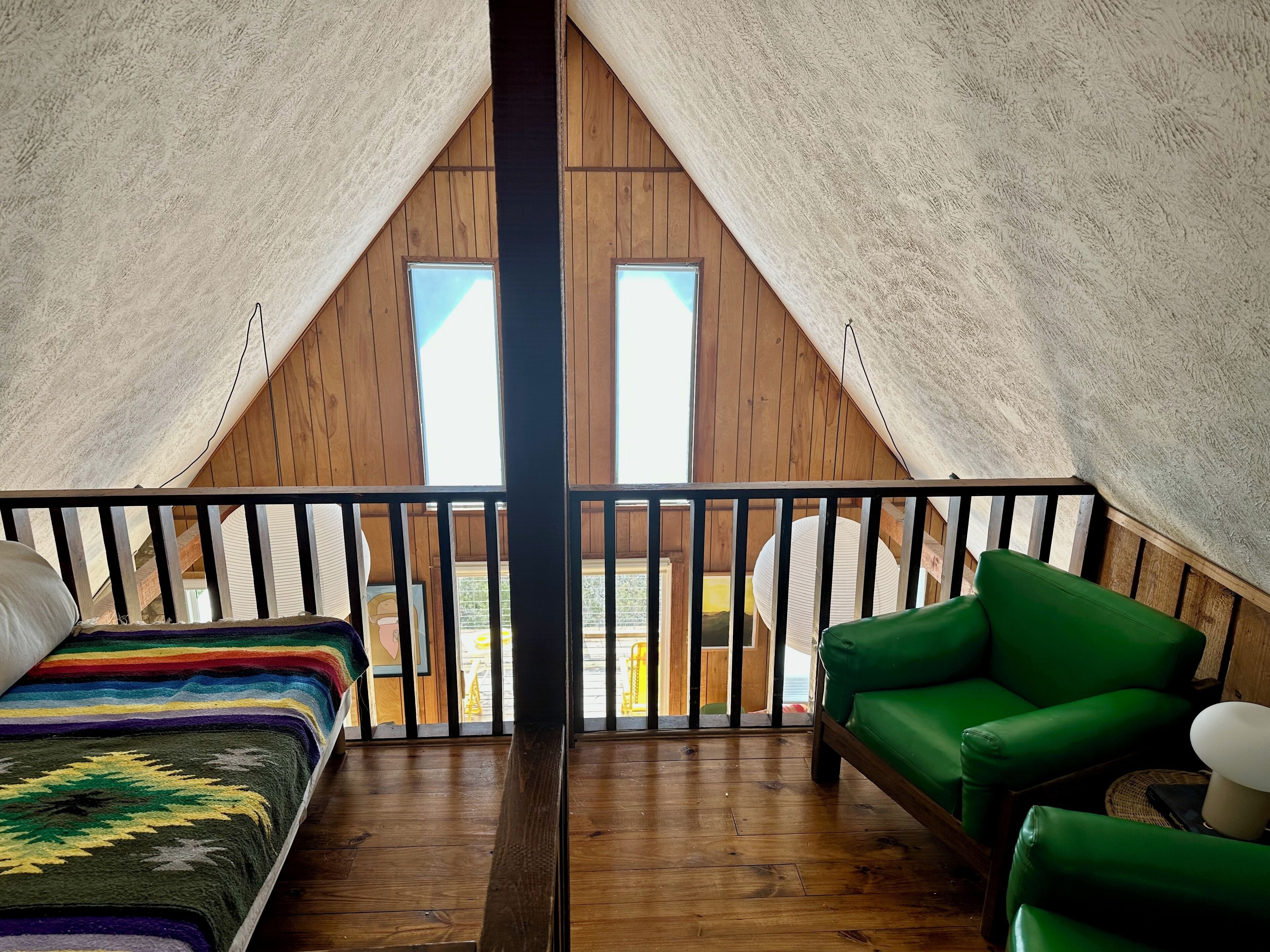 A cozy attic bedroom with wooden walls, a wooden floor, a green chair, a bed with a colorful blanket, and a balcony with a railing.