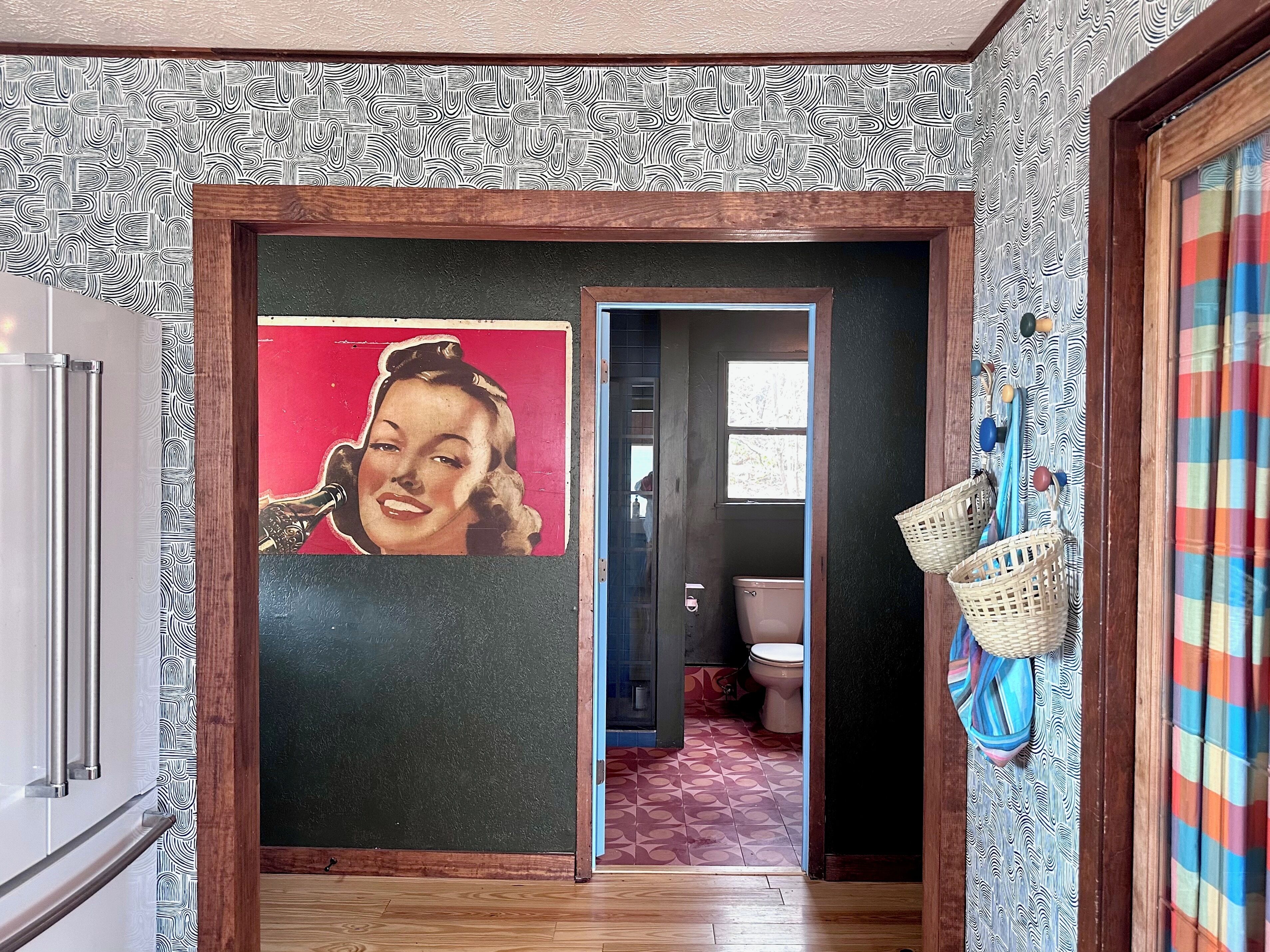 A hallway with patterned wallpaper, a door with a pop art print, a wooden floor, and a basket hanging on the wall.