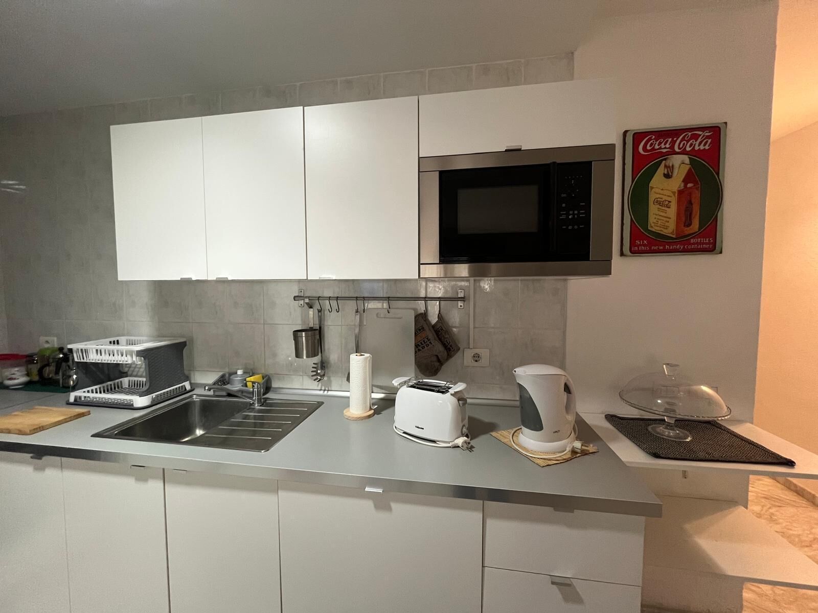 Una cocina moderna con armarios blancos, un lavamanos de acero inoxidable, un microondas, un tostador y una cafetera.