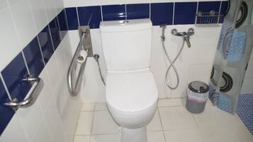 A bathroom with a white toilet, a handrail, a shower, and a trash bin.