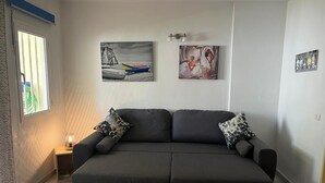 A modern living room with a dark grey sofa, two patterned throw pillows, and a small side table with a candle. There are three framed artworks on the wall above the sofa.