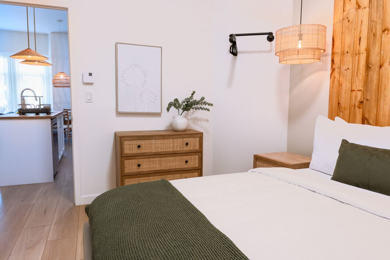 Quarto com cama, mesa de cabeceira, luminária na parede e vista para a cozinha.