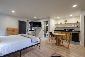 A modern bedroom with a bed, a bedside table, a TV, and a small kitchen area with a microwave, toaster, and a coffee machine.