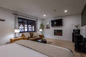 A modern living room with a large bed, a brown leather sofa, a flat-screen TV, a fireplace, and a view of the outdoors through a window.