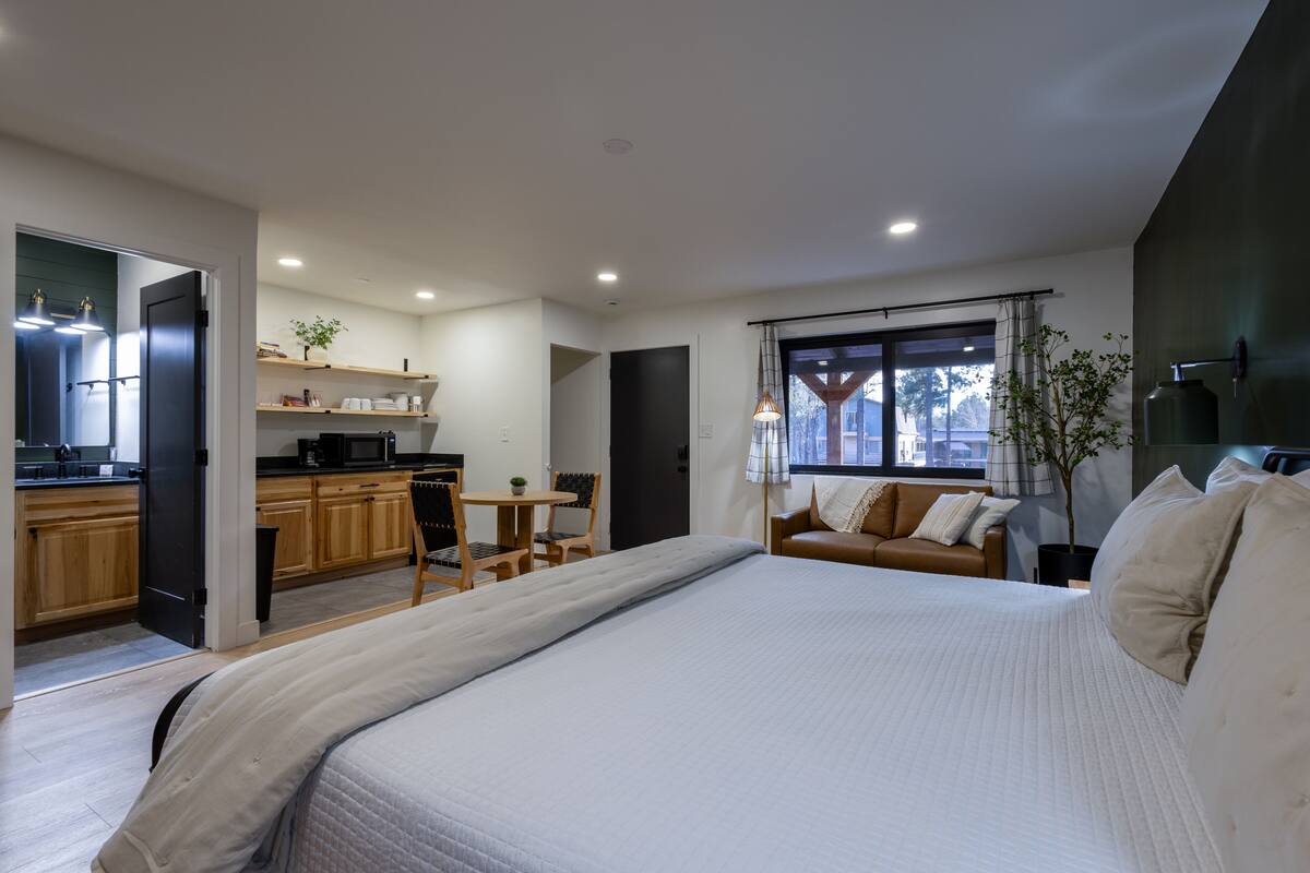 A modern bedroom with a large bed, a small desk, and a kitchenette in the background.