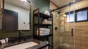 A bathroom with a large mirror, a sink, a glass shower enclosure, and shelves with towels and toiletries. - Timber Lodge (Pinetop-Lakeside)