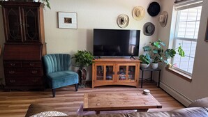 Una sala de estar con televisor, un sillón azul, una mesa de centro de madera, un sofá de cuero marrón, un aparador y varias plantas en macetas.