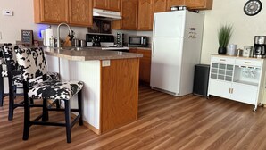 Una cocina con armarios de madera, un refrigerador blanco, un microondas y una cafetera. Hay un taburete de bar y un mueble pequeño con cristalería.