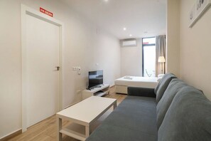 A compact hotel room with a bed, a sofa, a TV, a small table, and a door with a sign.