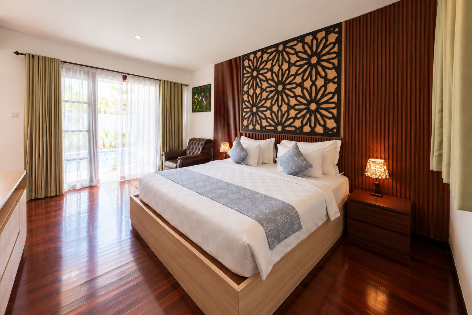A spacious bedroom with a large bed, a wooden headboard, a nightstand with a lamp, and a view of the outdoors through a sliding door.