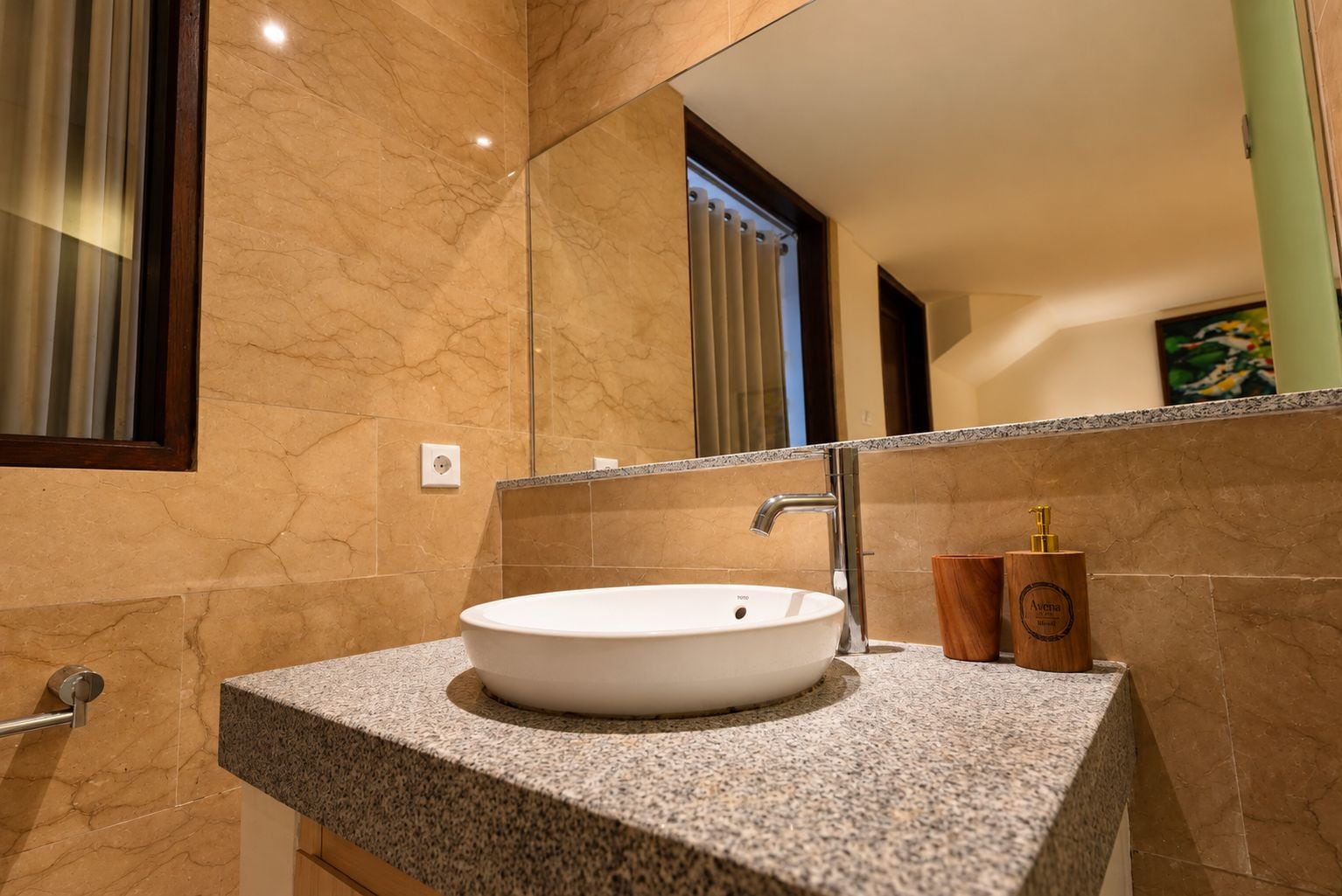 Un baño con lavamanos blanco, encimera de granito y un espejo que refleja una ventana con persianas.