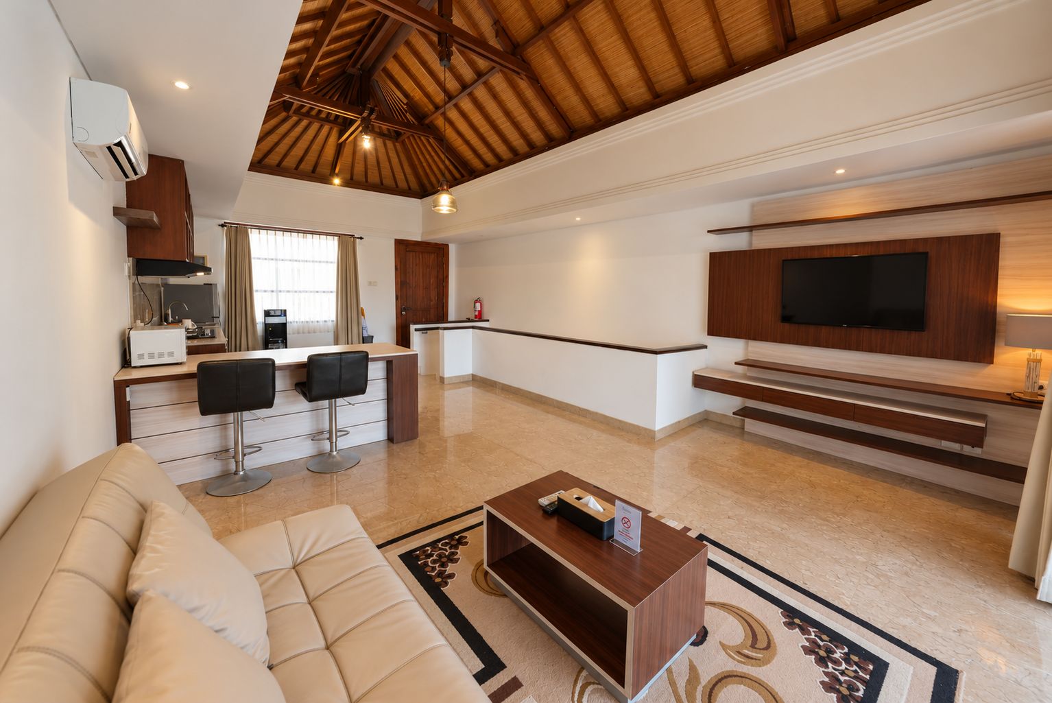A spacious living area with a sofa, a coffee table, a kitchen area with a dining table and chairs, a TV mounted on the wall, and a staircase leading to an upper level.
