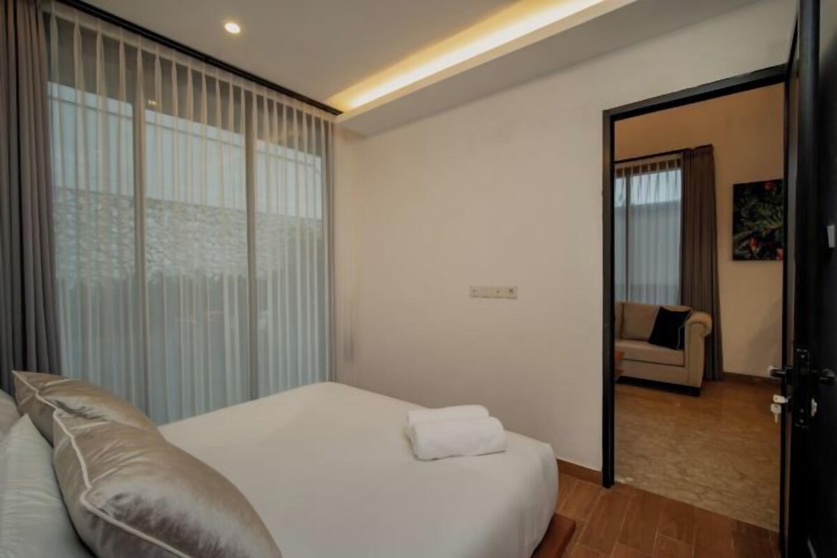 A bedroom with a bed, a window with blinds, a door leading to another room, and a light fixture on the ceiling.