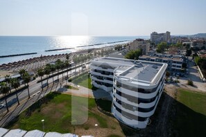 Um edifício com vários andares e terraço no telhado, ao lado de uma praia com guarda-sóis e palmeiras.