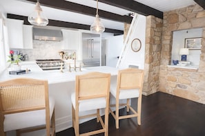 Una cocina moderna con armarios blancos, una isla grande y sillas de madera. Se observa una pared de piedra y una ventana con vistas.