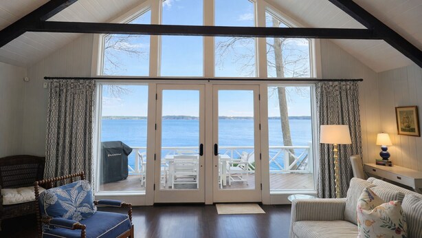 Une pièce spacieuse avec un plafond haut, de grandes fenêtres et une vue sur l’eau. Deux portes donnent sur une terrasse extérieure meublée de mobilier blanc.