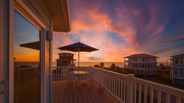 A wooden deck with a table and chairs, overlooking a beach and ocean during sunset. - Beachfront 5BR | Ocean Views, Elevator & Game Room (Palm Coast)