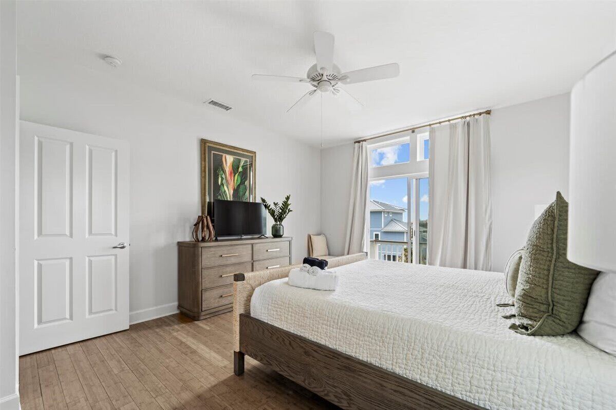 A bedroom with a bed, a ceiling fan, a dresser, a television, and a window with curtains.