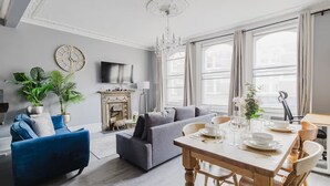 A modern living room with a fireplace, a dining table set for four, and a large window with curtains. - Elegant 3 Bedroom Fulham-Chelsea Home/ Rooftop Terrace/ Jacuzzi/ Sleeps 8 (London)