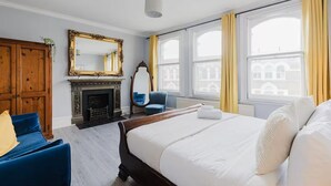 A hotel room with a large bed, a wooden wardrobe, a fireplace, a mirror, and large windows with curtains. - Elegant 3 Bedroom Fulham-Chelsea Home/ Rooftop Terrace/ Jacuzzi/ Sleeps 8 (London)