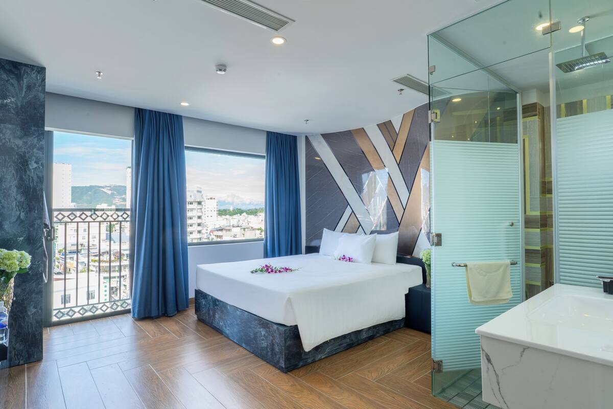 A modern hotel room with a large bed, a bathroom with a large mirror, and a balcony with a view of the city.