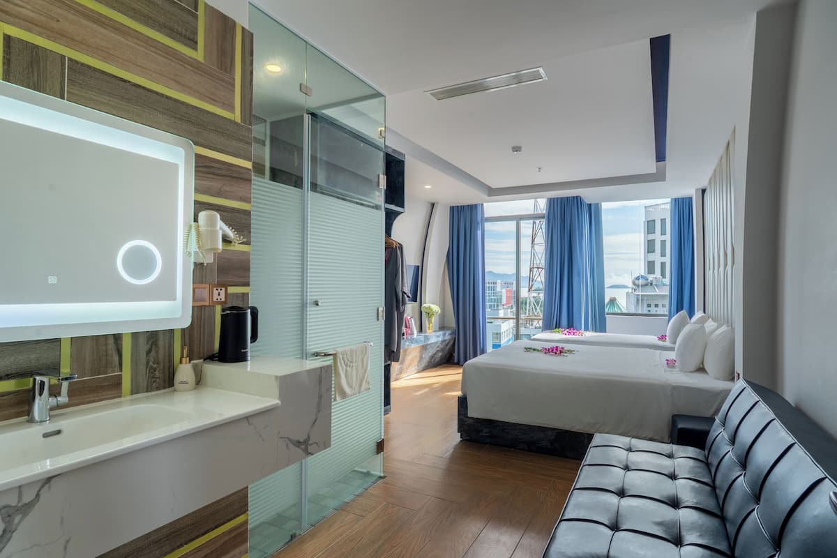 A modern hotel room with a large bed, a sofa, a vanity with a mirror, and a view of the city through large windows.