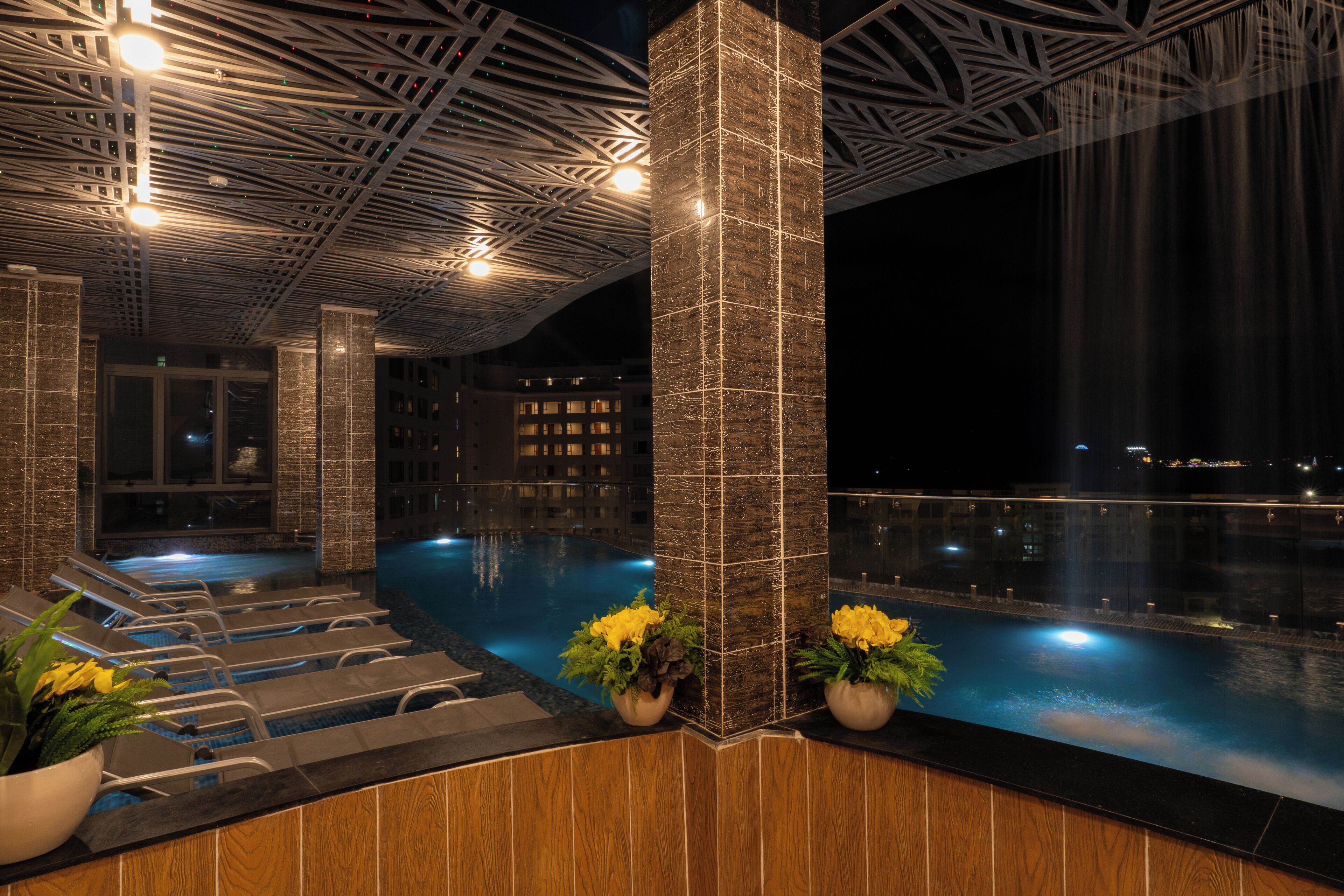Ein beheiztes Hallenbad mit modernem Deckenbau, ein Poolbereich mit Liegestühlen und Blick auf die nächtliche Skyline.