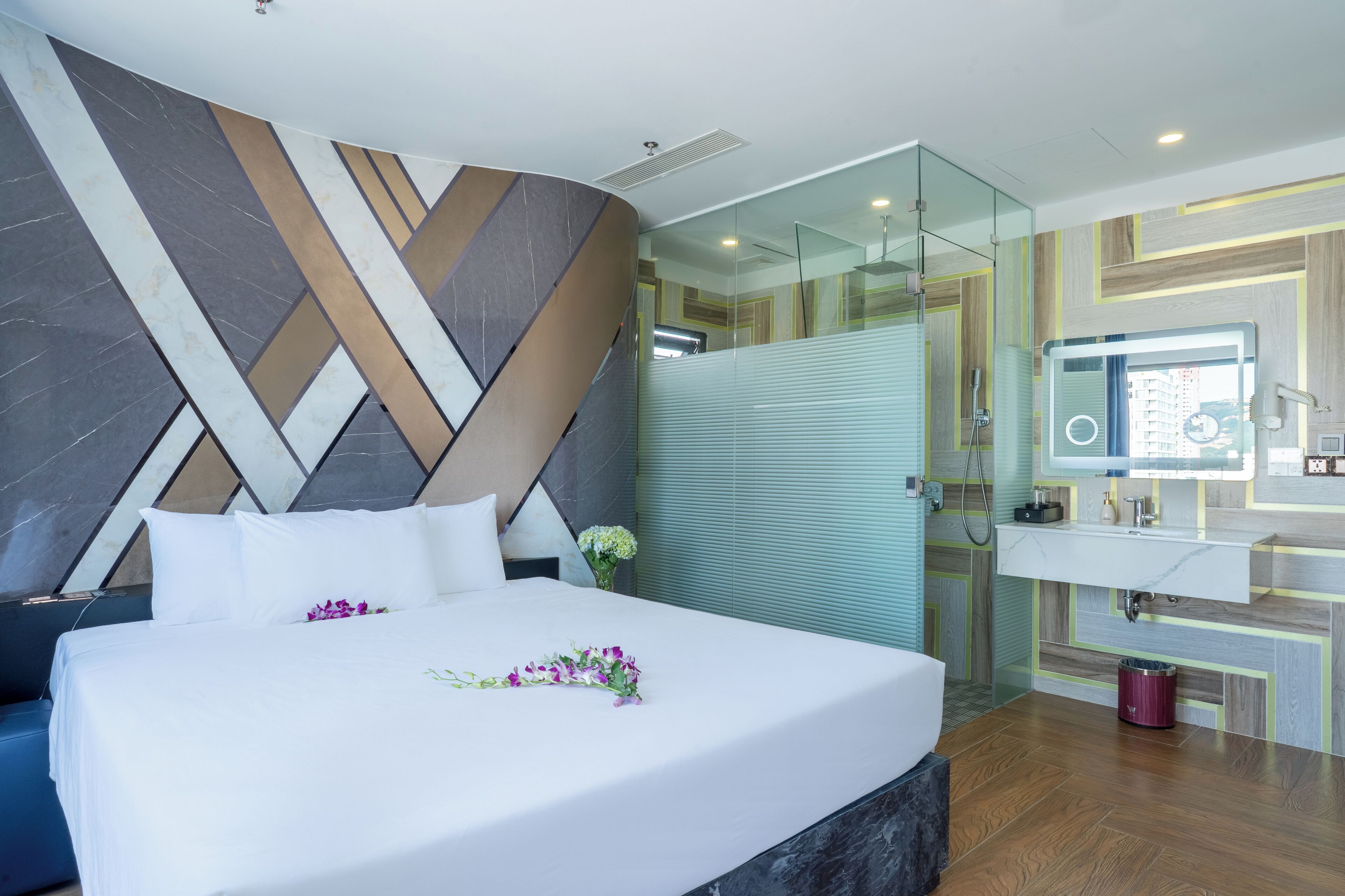 Una habitación de hotel moderna con una cama grande, cabina de ducha de vidrio, baño con espejo y lavamanos, y un diseño decorativo en la pared.