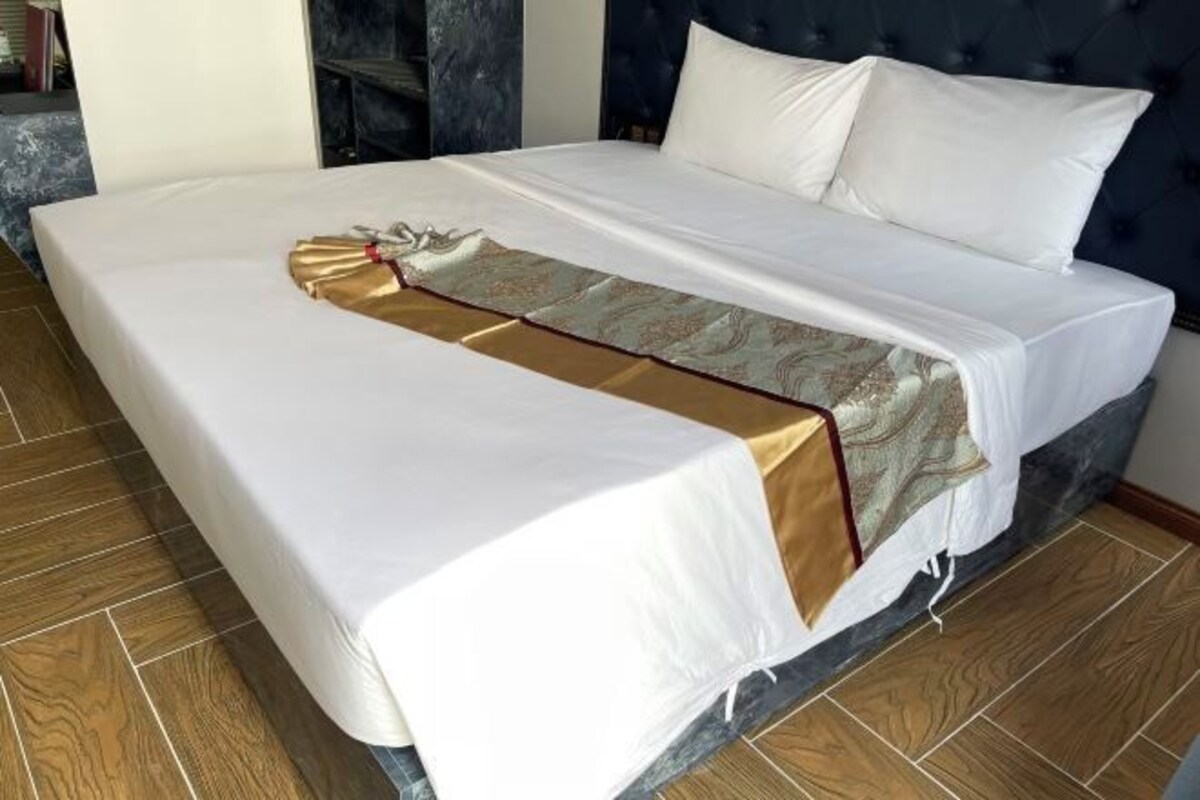 A neatly made bed with a white and gold bedspread, a dark headboard, and a wooden floor.