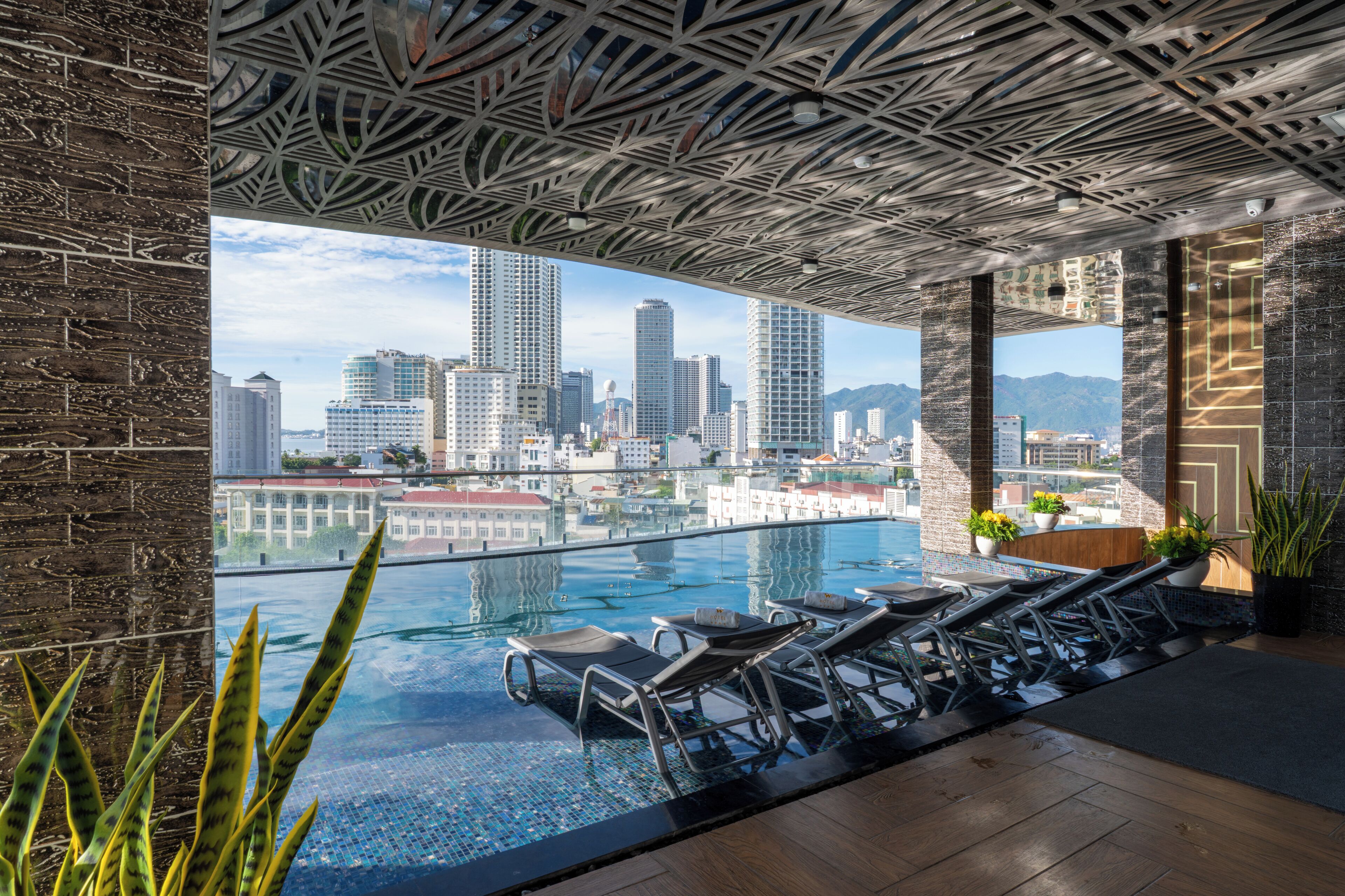 A rooftop infinity pool with lounge chairs, a city view, and a patterned ceiling.