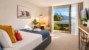A hotel room with a bed, a desk, a chair, a TV, a balcony with a view of mountains and a lake. - Lake View Lodge (Queenstown)