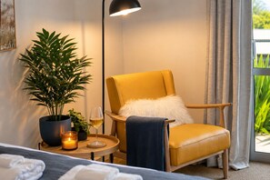 A room with a yellow armchair, a potted plant, a candle, and a glass of wine on a small table.