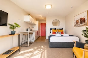 A compact hotel room with a bed, bedside table, and a desk with a chair. There is a small kitchenette and a mirror on the wall. - Lake View Lodge (Queenstown)