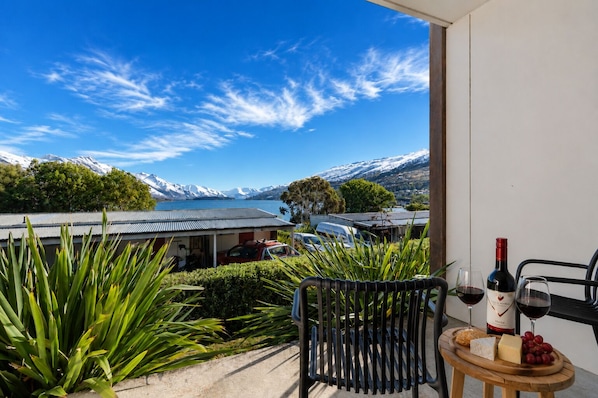 A balcony with a view of snow-capped mountains, a table with a bottle of wine, cheese, and grapes, and a chair.