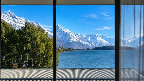 A view of a lake with snow-capped mountains in the background, a wooden deck, and a building with a sloped roof. - Lake View Lodge (Queenstown)