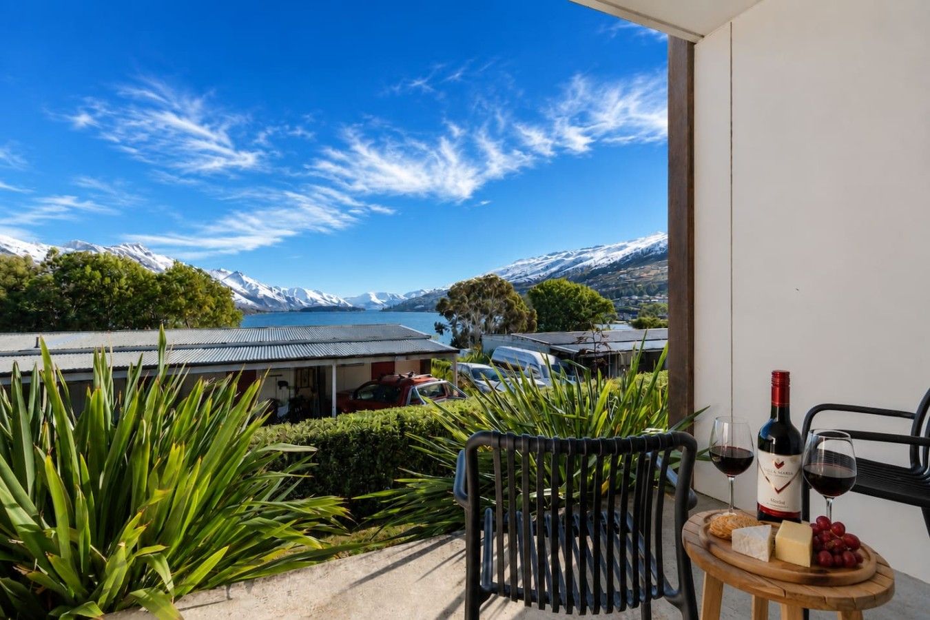 Een balkon met uitzicht op een meer en bergen, een tafel met een fles wijn, glazen, kaas en druiven.