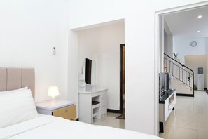 A modern hotel room with a bed, bedside table, lamp, and a small desk with a mirror and TV. - Villa Normandie (Cimahi)