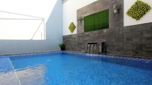 An indoor swimming pool with a tiled floor, a wall-mounted water feature, and decorative plants. - Villa Normandie (Cimahi)
