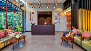 A reception area with a receptionist, two sofas, a wooden reception desk, and a large window with a view of greenery. - Vbrings Oak Village (Devikolam)