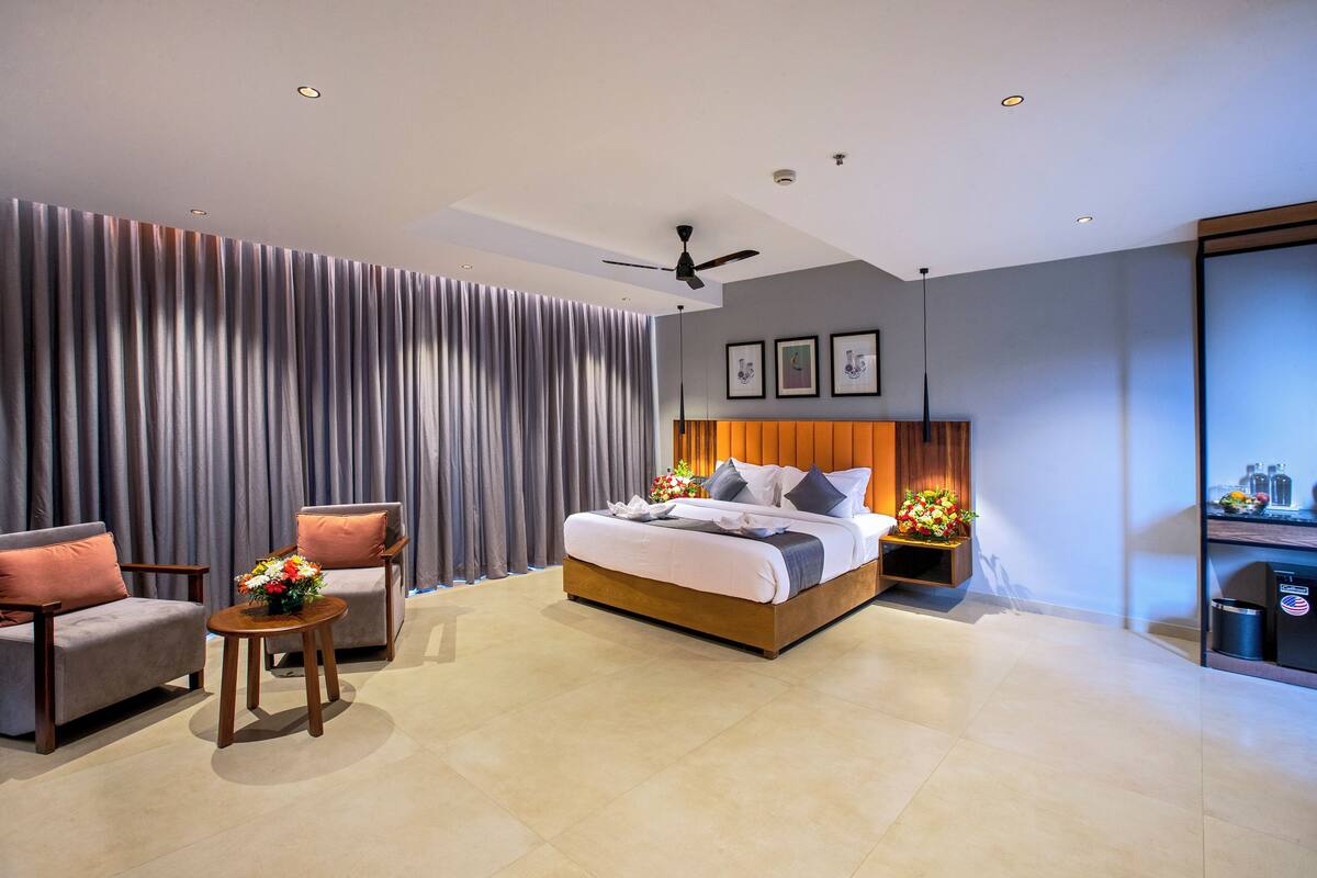 A modern hotel room with a large bed, two armchairs, a small table with a vase, and a minibar.