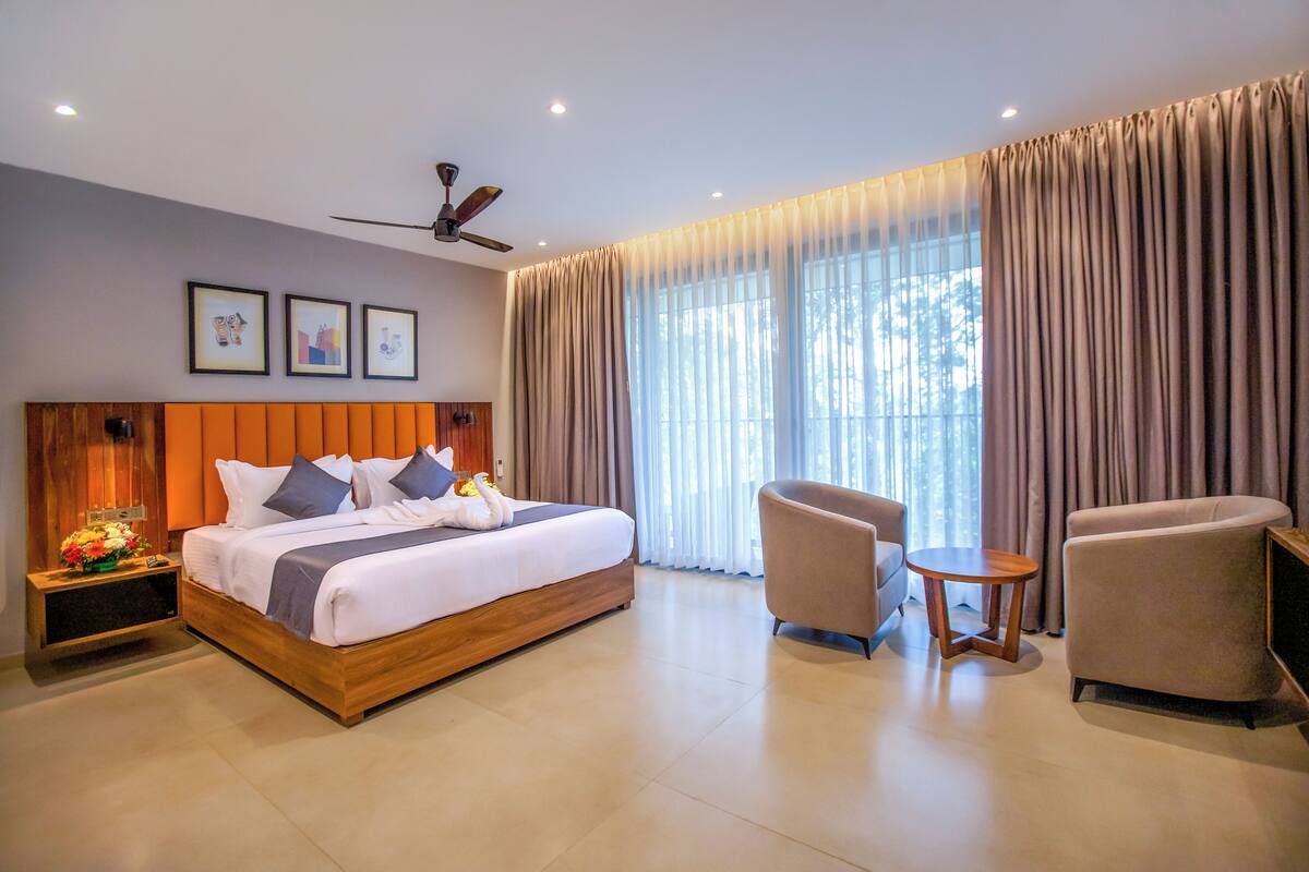 A modern hotel room with a large bed, a sitting area with a chair and a small table, a ceiling fan, and a large window with curtains.