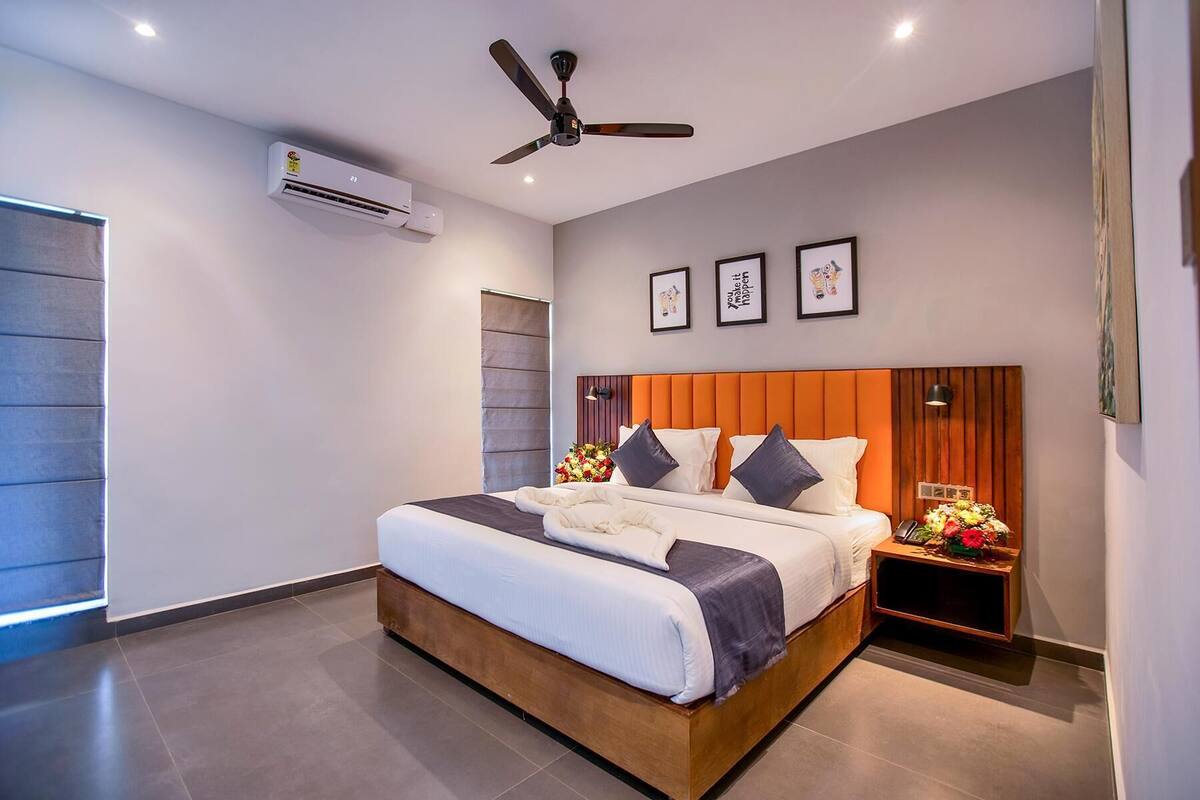 A modern hotel room with a large bed, a ceiling fan, an air conditioning unit, and a nightstand with a lamp and flowers.