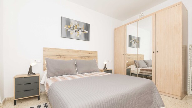 Een slaapkamer met een houten hoofdbord, een groot bed met een grijze quilt, een nachtkastje met een lamp, een kledingkast en een lijst met een afbeelding aan de muur.