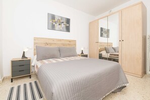 Une chambre à coucher avec une tête de lit en bois, un grand lit recouvert d’une couette grise, une table de chevet avec une lampe, une armoire et un tableau encadré accroché au mur.