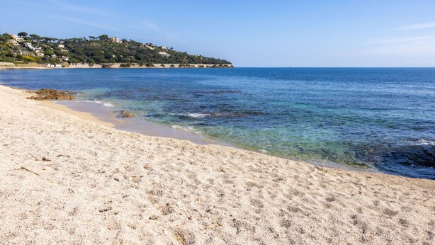 Ein Strand mit klarem Wasser, sandigem Ufer und Gebäuden, die sich auf einem Hügel im Hintergrund erheben.